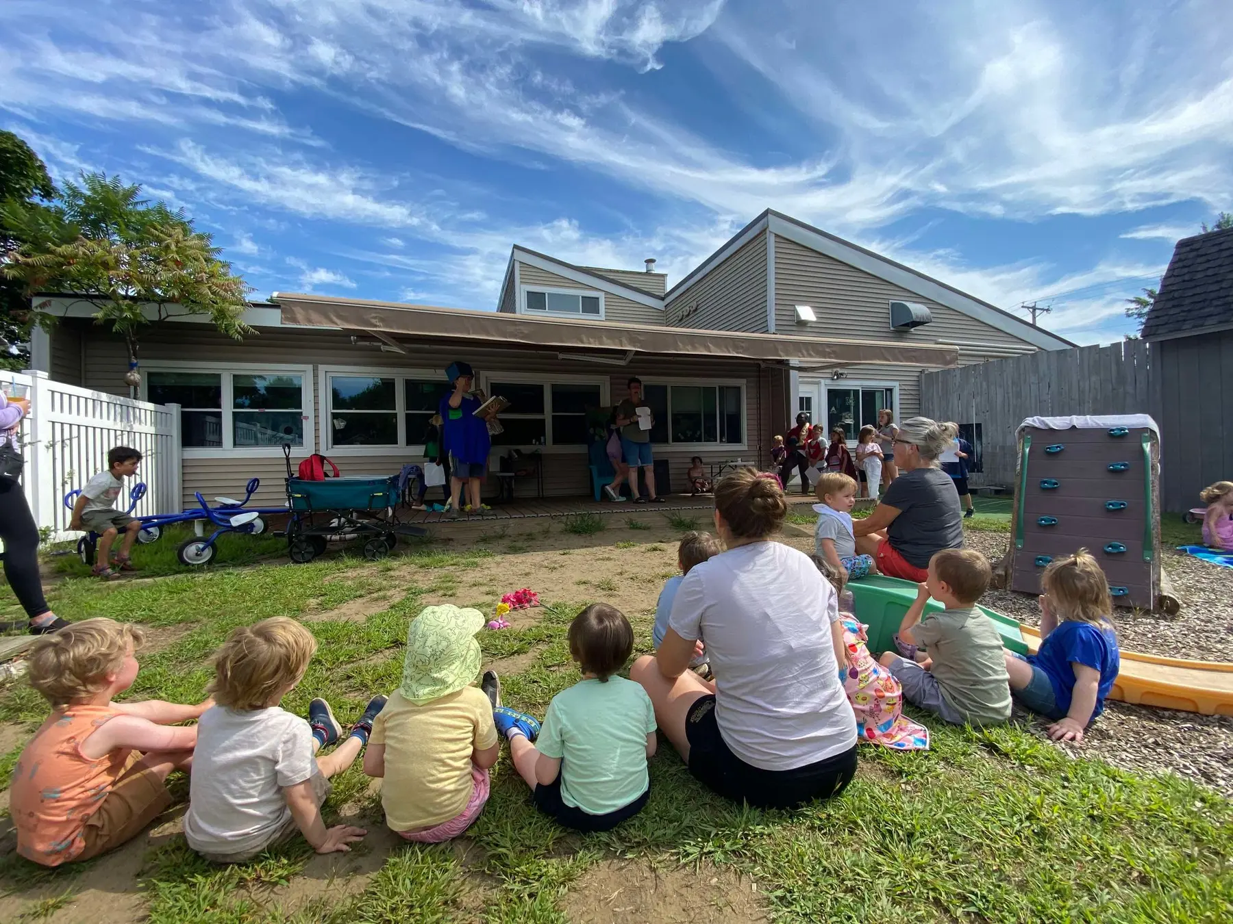 Children and adults sit on grass and gravel in a backyard while a person stands under an awning reading from a book to the group.