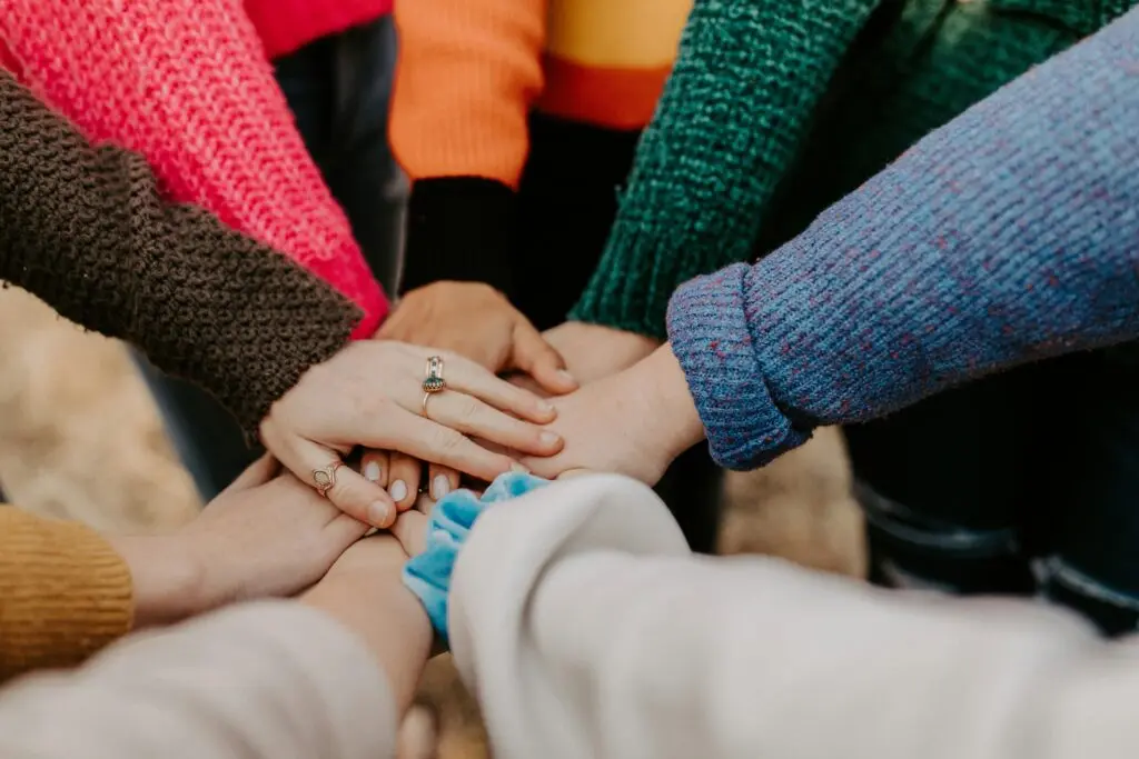 Several people in colorful knit sweaters stand in a circle with their hands stacked together in the center.