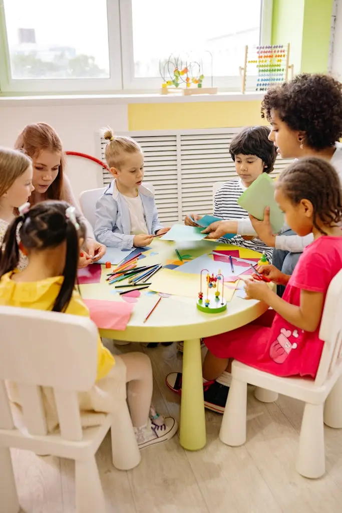 Children and adults sit around a low table drawing with colored pencils and handling paper shapes in a bright room.