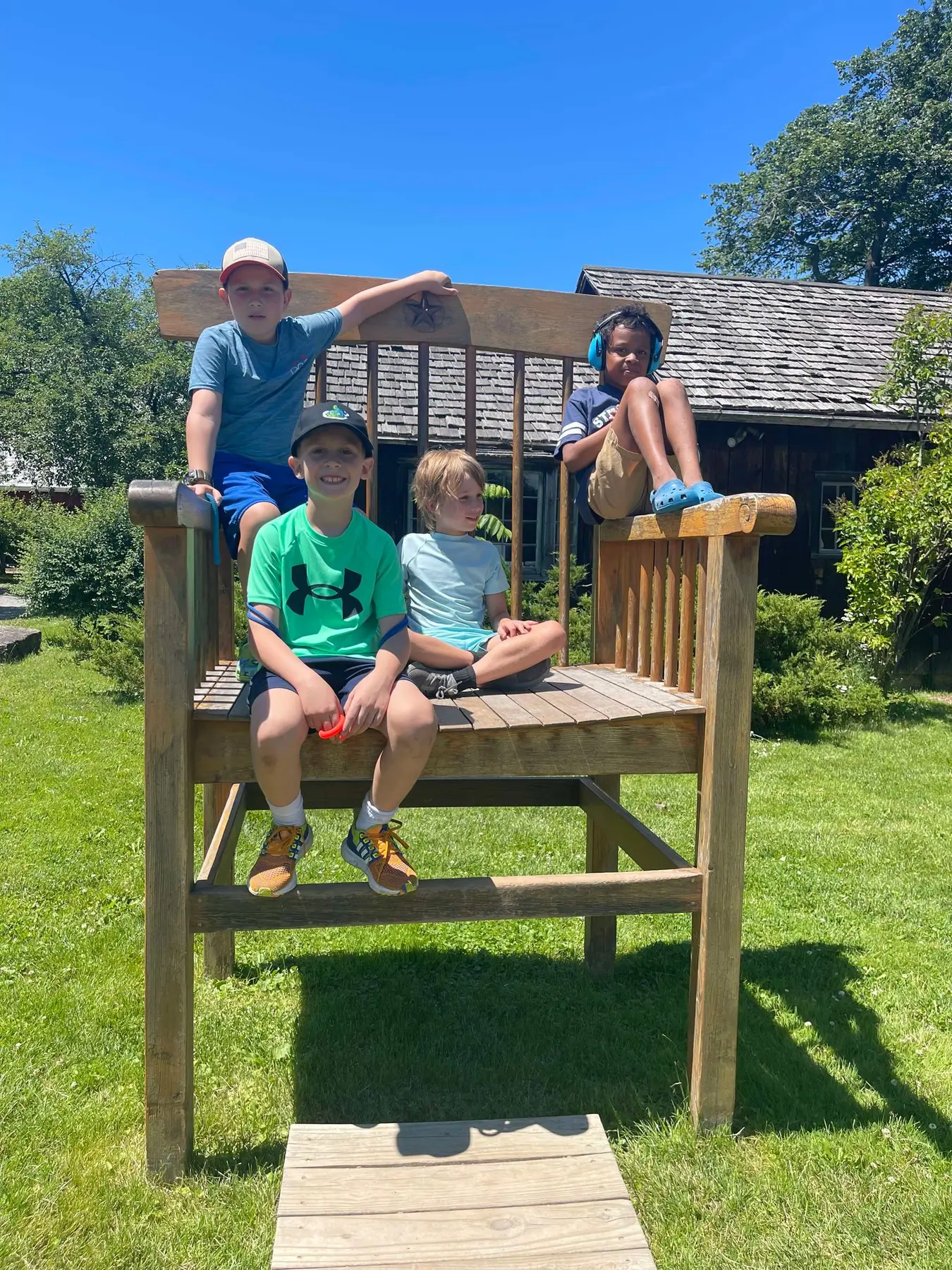 Four children sit and pose on a large wooden chair structure on a grassy lawn in front of a building with a shingled roof.