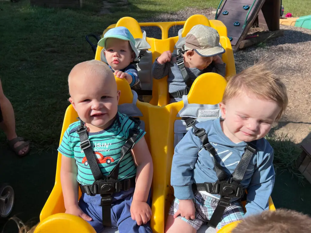 Four young children sit strapped into a yellow multi-seat stroller outdoors, with two facing forward in the front and two facing forward in the back.