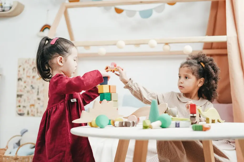 Two young girls stand at a small white table, one in a red dress placing a toy block while the other in a beige dress reaches for a toy, surrounded by wooden toys and a play tent.