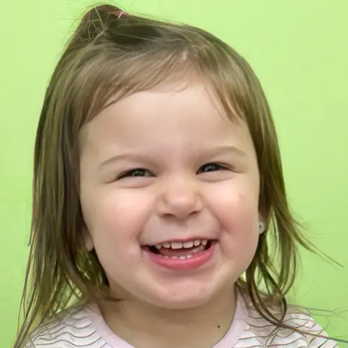 A smiling girl with a small ponytail and a striped shirt looks directly at the camera against a solid green background.