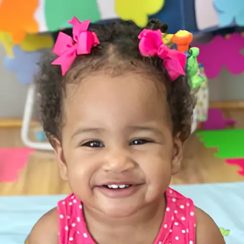 A smiling toddler wearing a pink polka-dot top and two pink hair bows sits in front of colorful wall decorations.