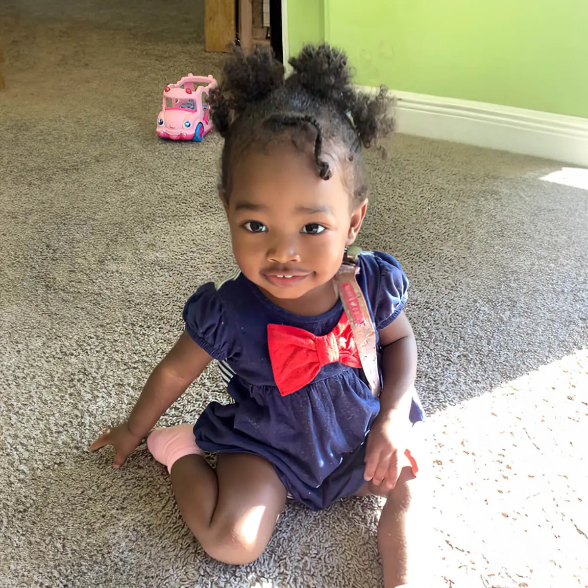 A young girl with two hair buns sits on a carpeted floor wearing a navy blue dress with a red bow and a pink sock, with a pink toy car behind her.
