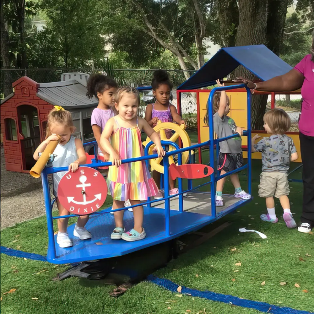 Several young children stand and play on a blue play structure with a ship wheel and red anchor sign, while an adult stands to the right on green artificial turf.