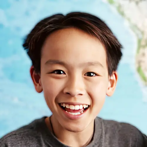 A smiling child with short dark hair wearing a gray shirt is shown against a blurred blue and green background.