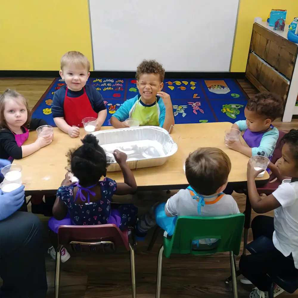 Several young children sit around a table with clear cups and a foil tray, while an adult in blue gloves holds a cup in the foreground.