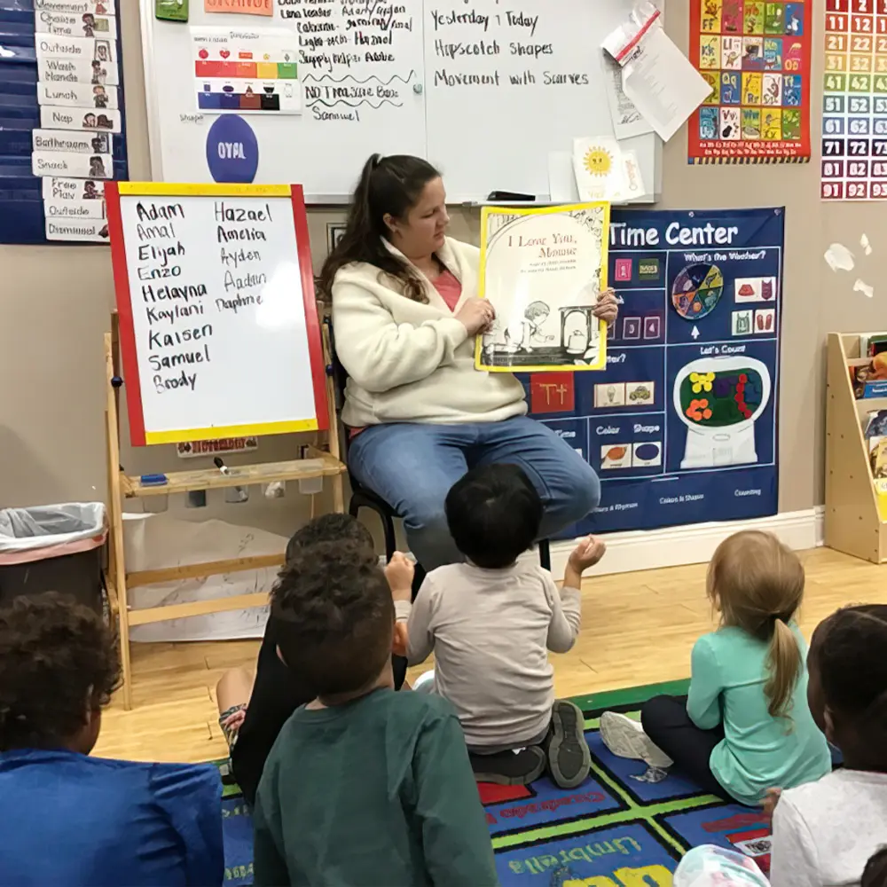 A woman sits on a chair holding a book open while six children sit on a colorful rug facing her in a classroom with educational posters and a whiteboard.