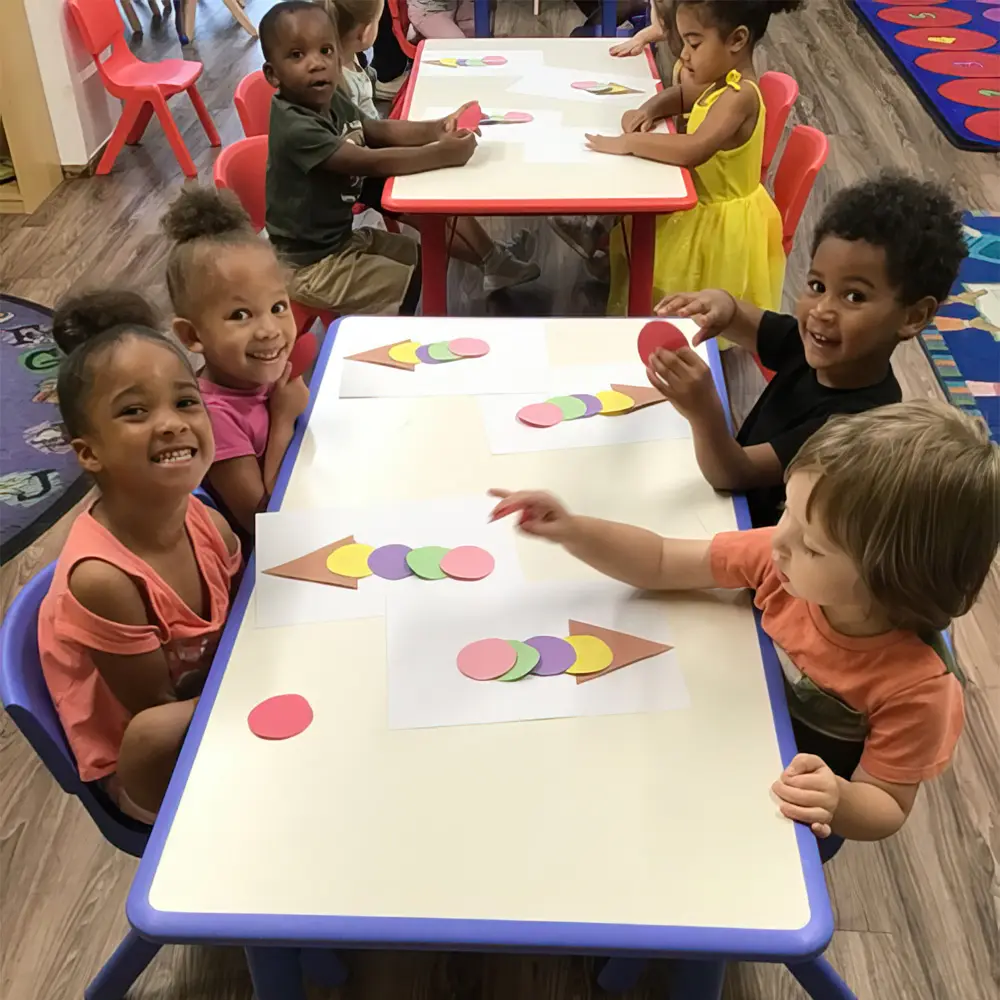 Children sit at two tables arranging colored paper circles and triangles on white paper while looking toward the camera.