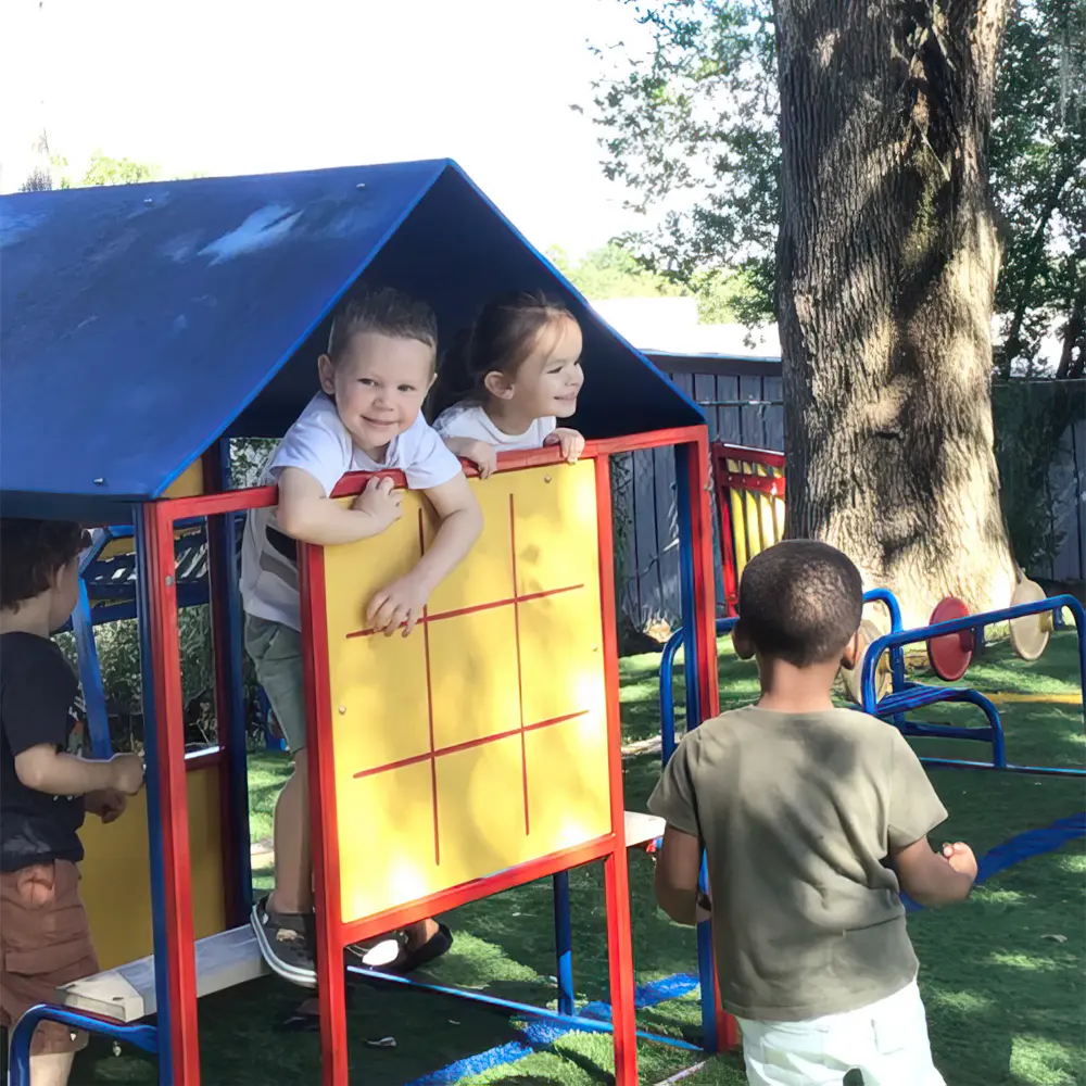 Four children play on a blue and red outdoor play structure with a yellow tic-tac-toe panel, while two children lean over the panel and two others stand nearby on green surfacing.