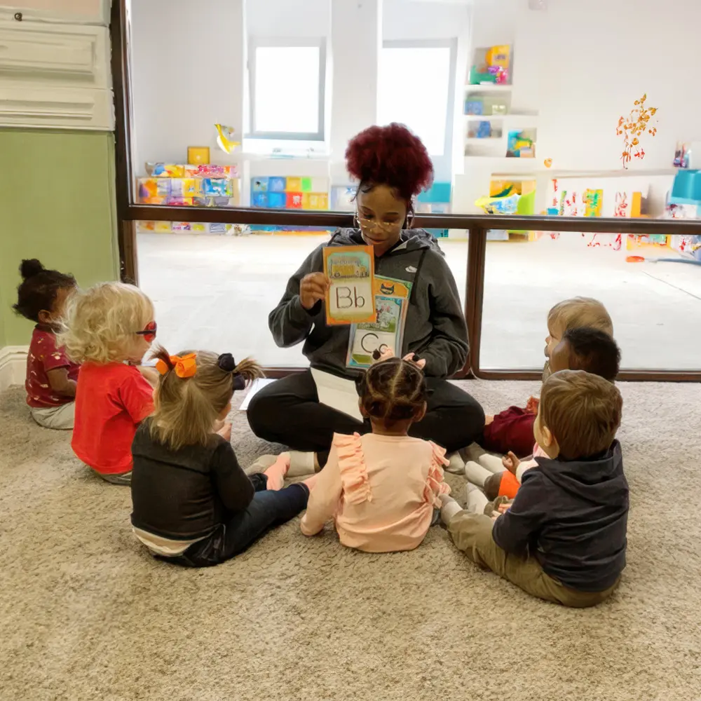 An adult sits on a carpeted floor holding alphabet cards while seven young children sit in a semicircle facing her in a classroom.
