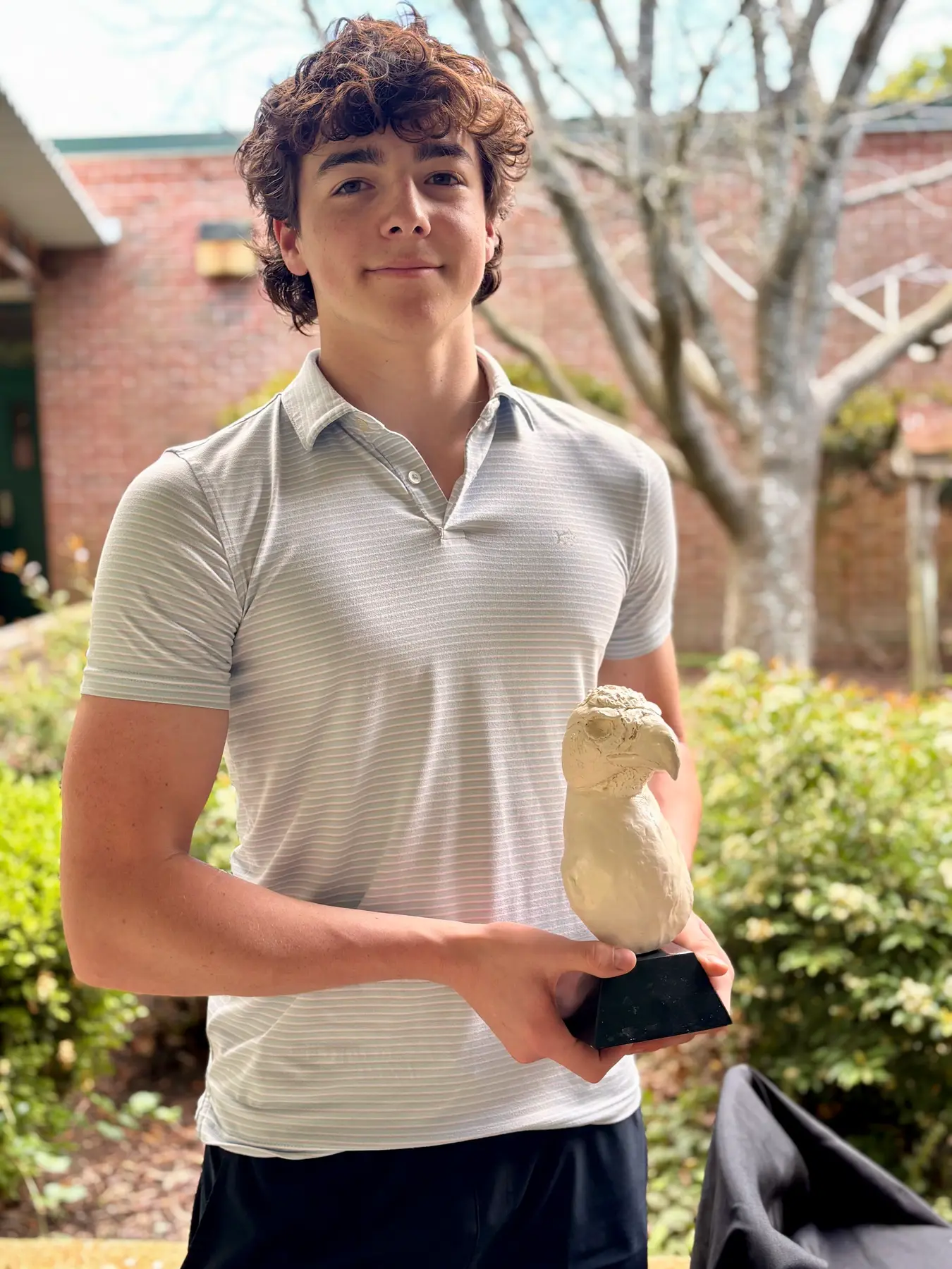 A young person in a striped polo shirt stands outdoors holding a white bird sculpture on a black base.