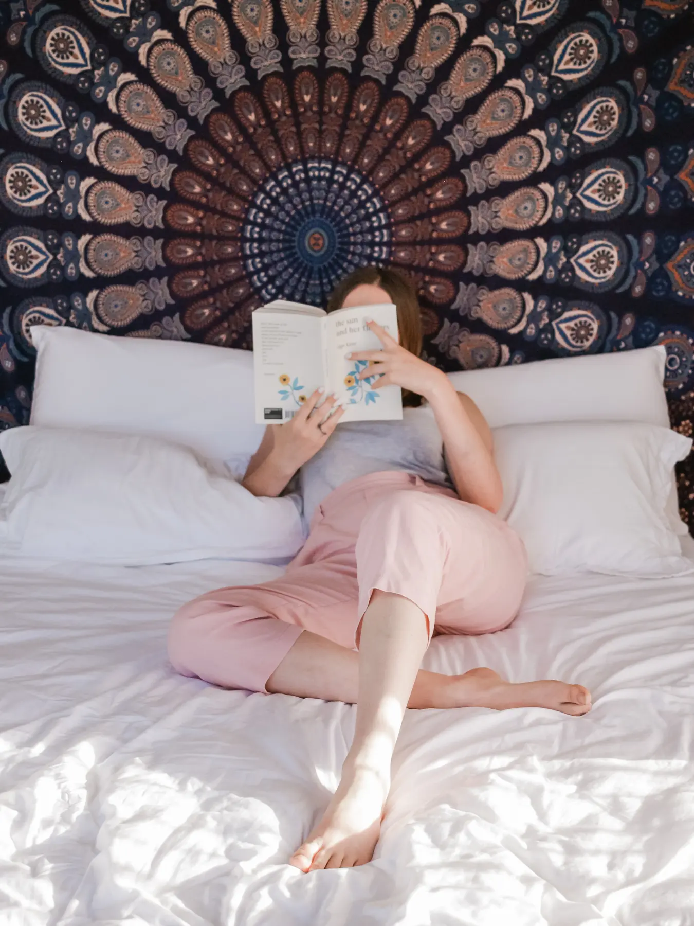 A person sits cross-legged on a white bed holding an open book in front of their face, with a large patterned tapestry hanging on the wall behind them.
