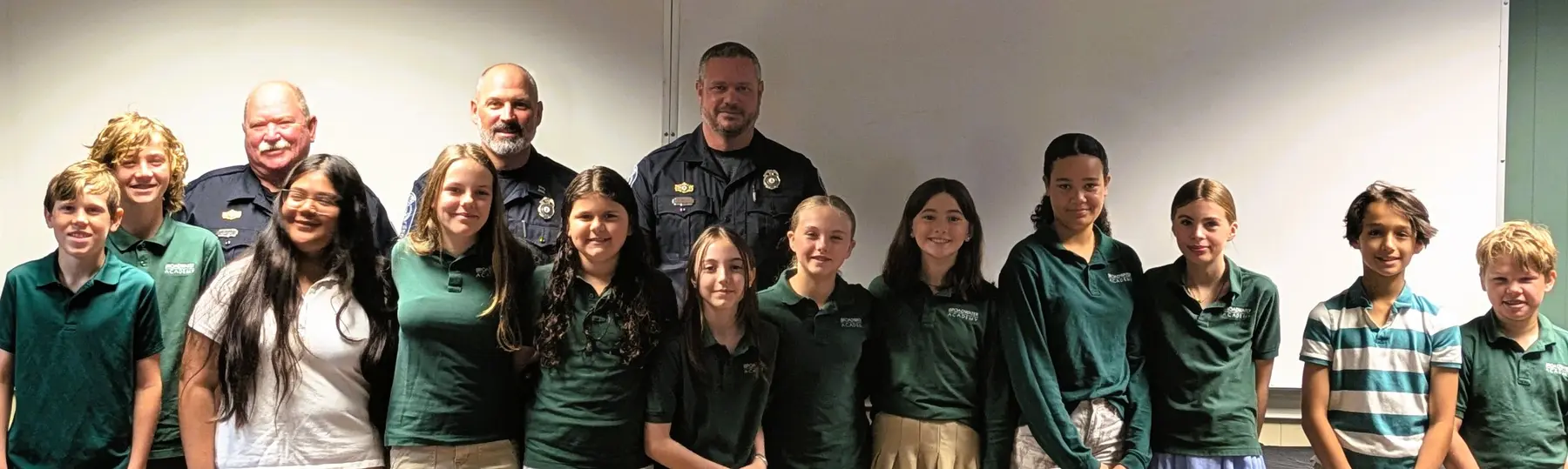 A group of children and adults in green shirts and uniforms stand together facing the camera in front of a white wall.