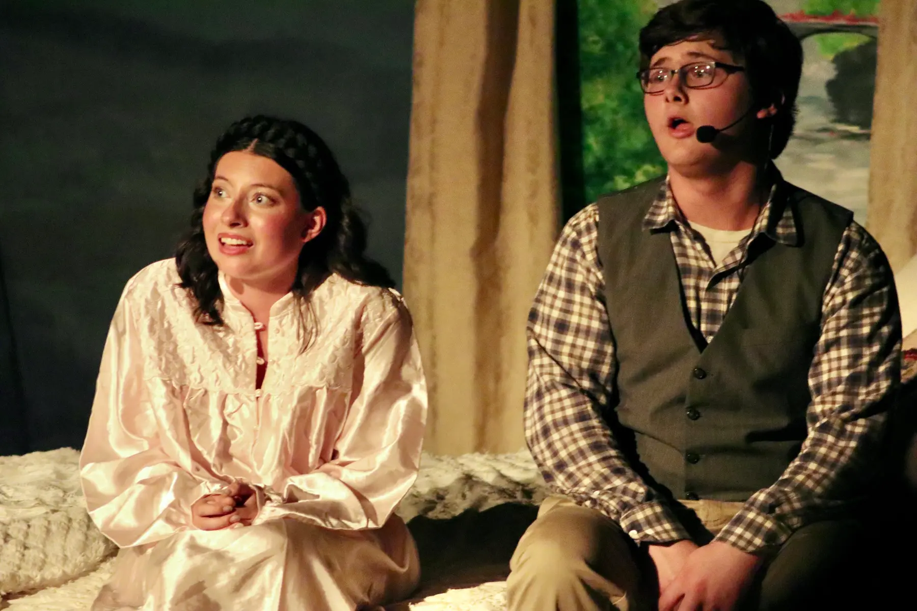 A woman in a shiny white nightgown and a man in a plaid shirt and vest sit on a textured surface, both with open mouths as if singing or speaking.