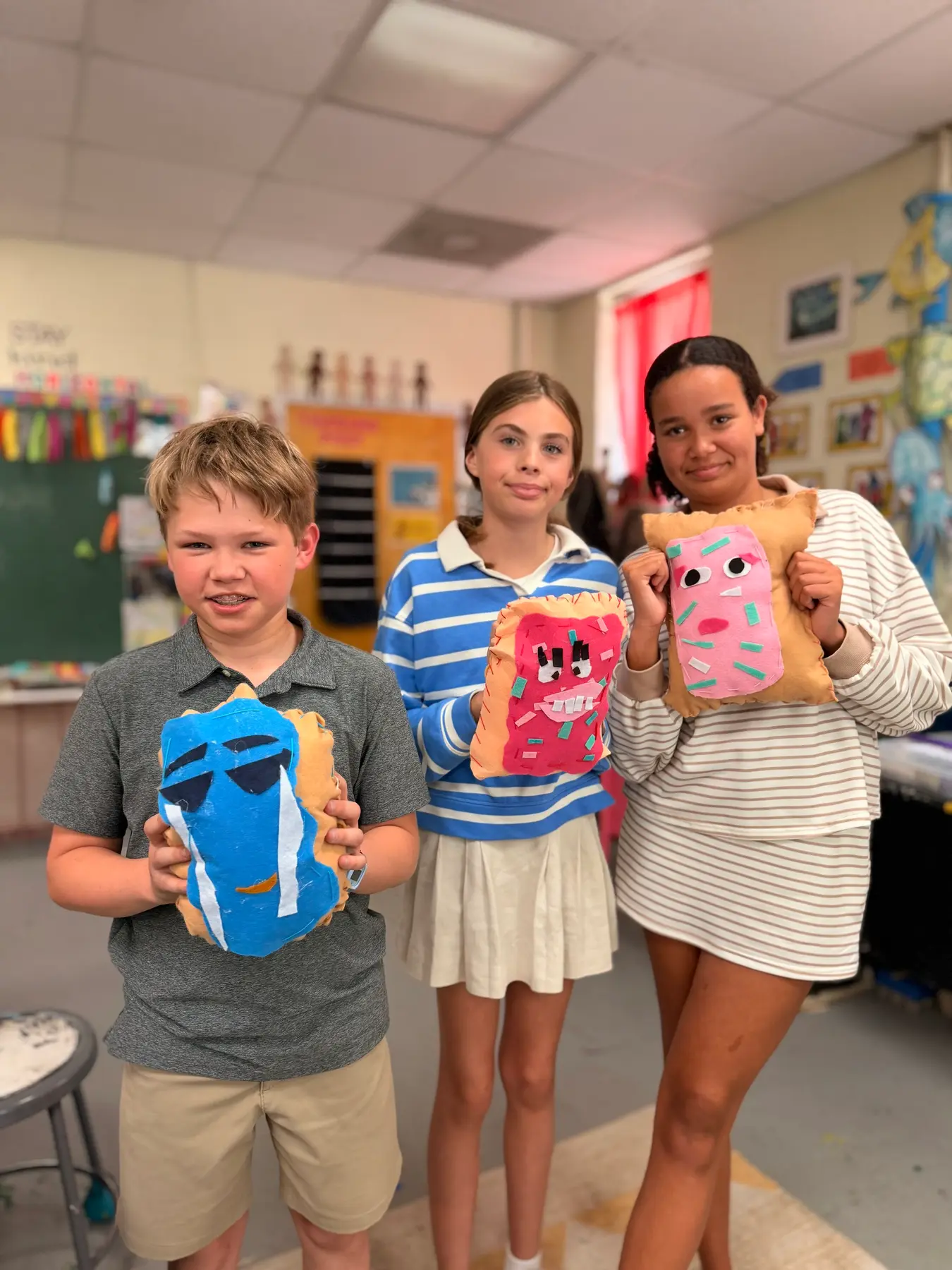 Three children stand in a classroom holding stuffed fabric pillows decorated with painted faces and paper shapes.