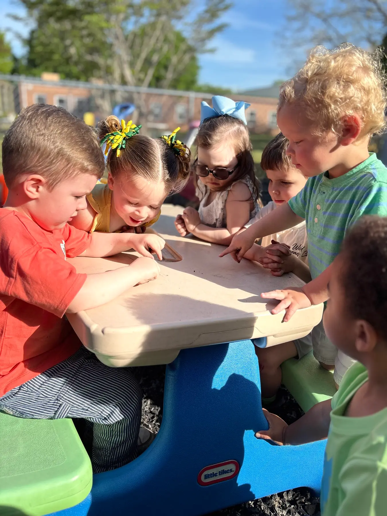 Six children gather around a blue and beige plastic table outdoors, looking down and pointing at the surface.