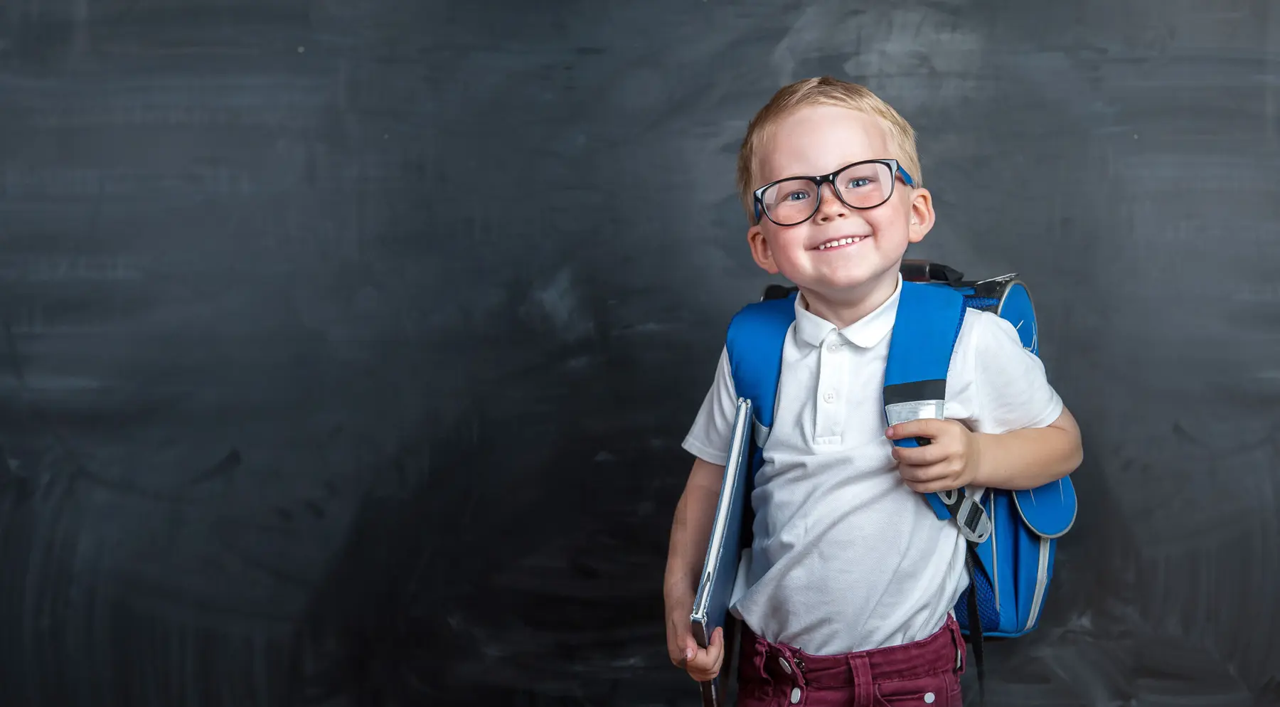 A smiling boy wearing glasses, a white polo shirt, and a blue backpack holds a folder while standing in front of a dark chalkboard.