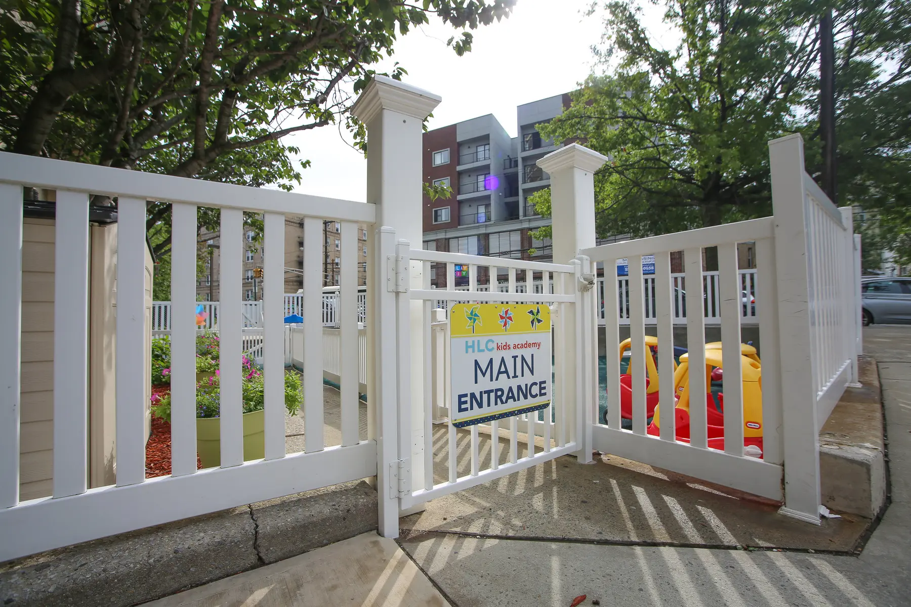 A white fence with an open gate displaying a sign reading 'MAIN ENTRANCE' stands on a sidewalk in front of a building and trees.