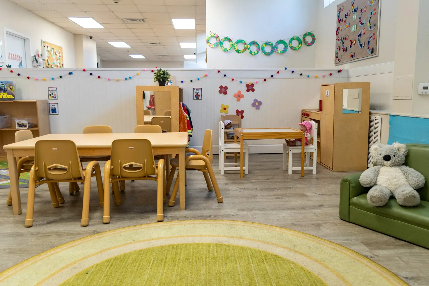 A childcare classroom with a large wooden table and chairs, a small table with chairs, a green sofa with a stuffed bear, and a circular rug on the floor.