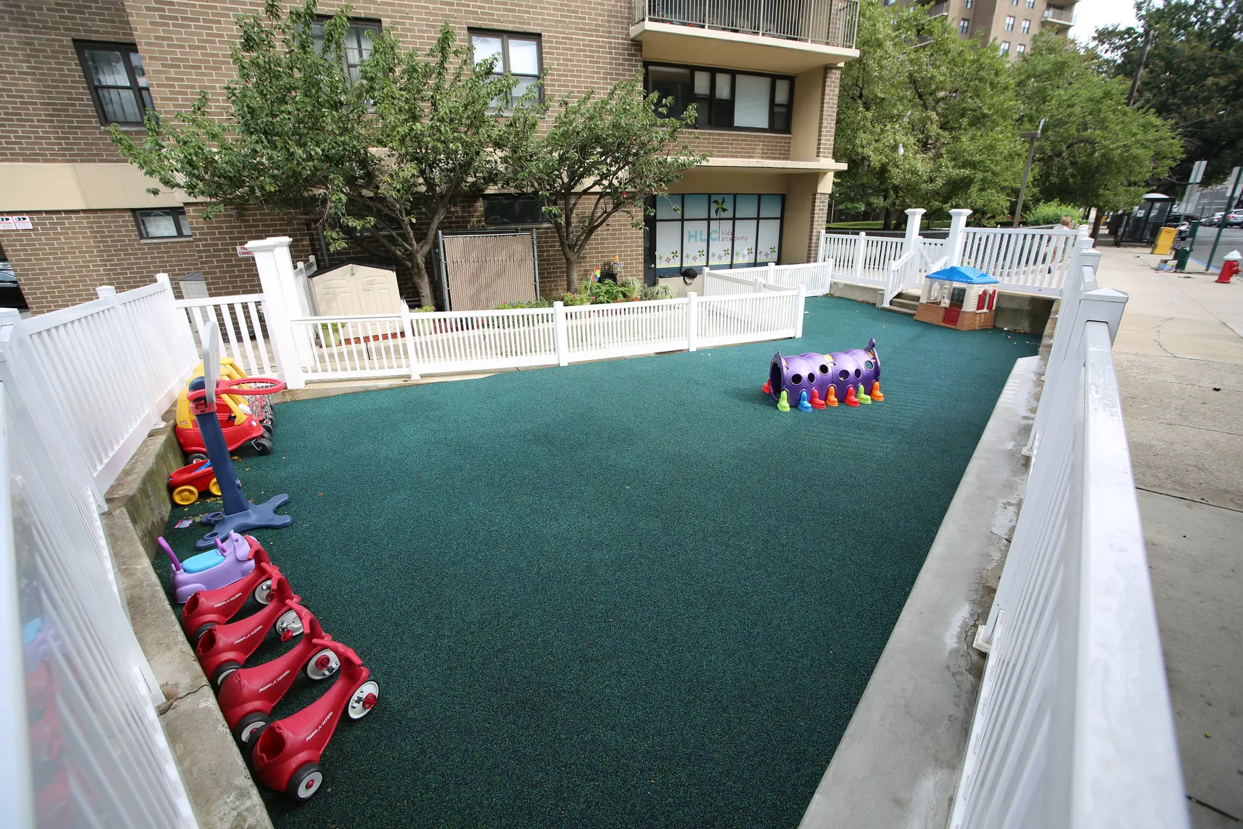 A fenced outdoor play area with green surfacing contains a basketball hoop, several red ride-on toys, a purple tunnel, and a small playhouse beside a brick apartment building.