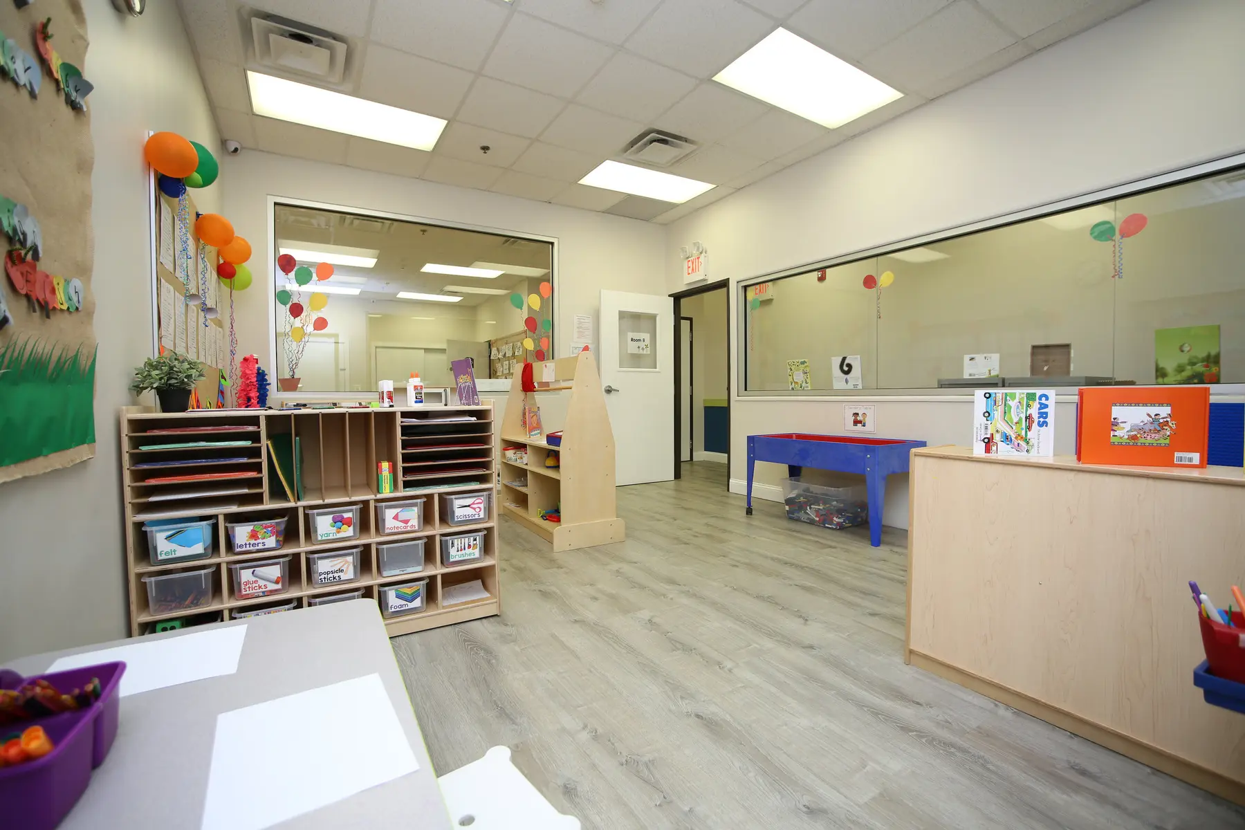 An empty preschool classroom with wooden storage shelves, a blue table, wall decorations, and large windows.