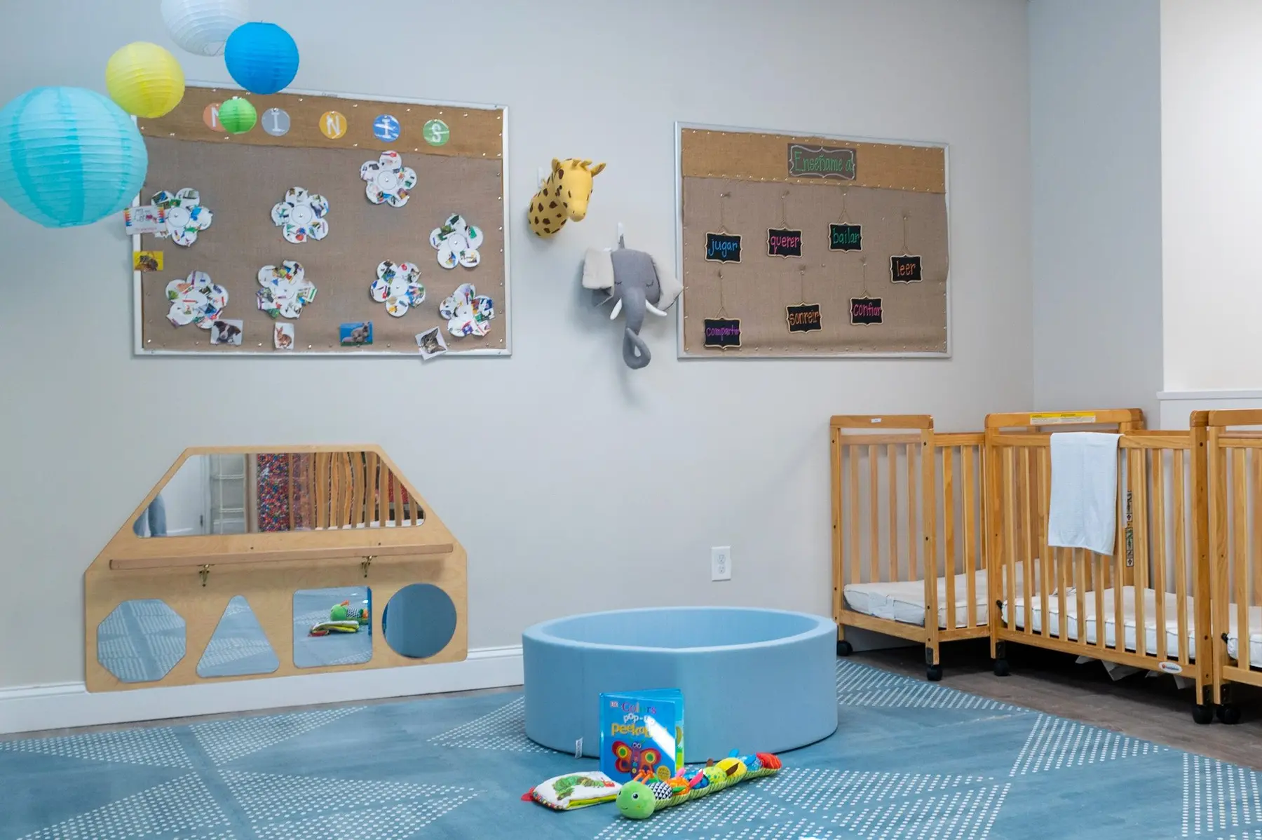 A childcare room with two wooden cribs, a wall-mounted mirror, bulletin boards, hanging paper lanterns, and a blue play mat holding a small tub and toys.