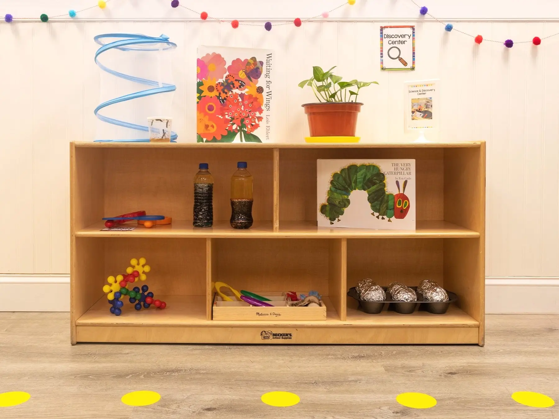 A wooden storage shelf holds books, bottles, toys, and plants in a classroom setting with a polka-dot rug on the floor.
