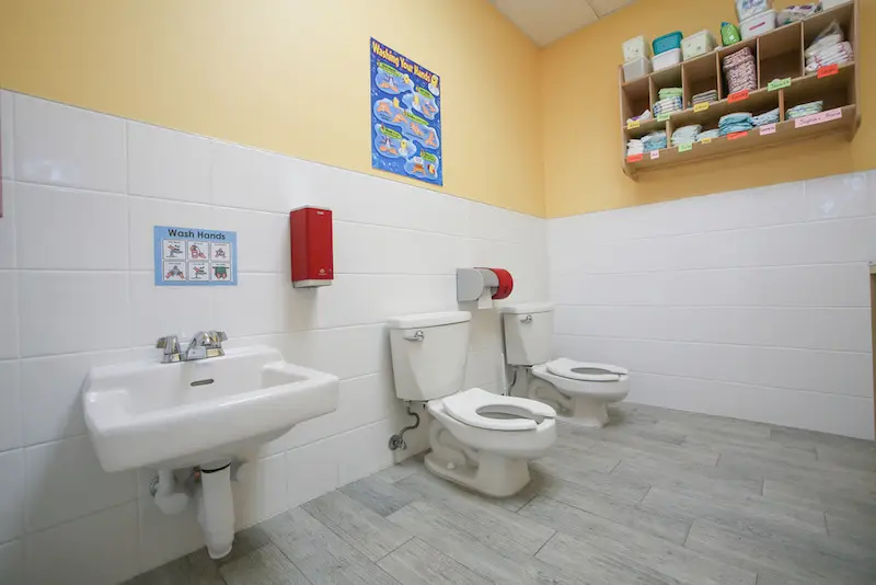 A childcare bathroom with a small sink, two child-sized toilets, a red paper towel dispenser, a handwashing poster, and a wall shelf holding diapers and wipes.