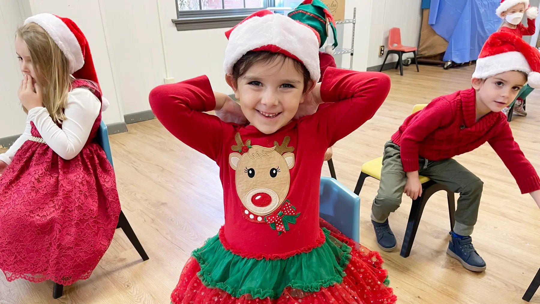 Children in Santa hats and red holiday clothing sit on chairs in a classroom, with one girl in the foreground posing with her hands behind her head.