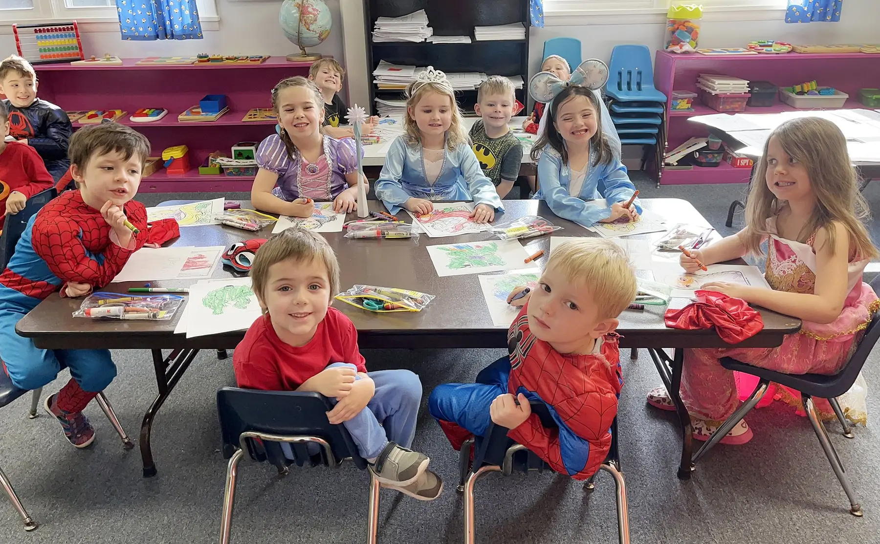Children in costumes and casual clothes sit and stand around a table coloring pictures with crayons in a classroom.