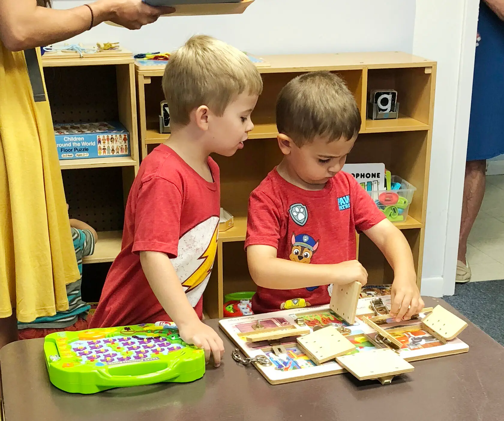 Two boys in red shirts stand at a table, one watching as the other manipulates wooden puzzle pieces on a board, with a green toy and an adult's arm visible nearby.