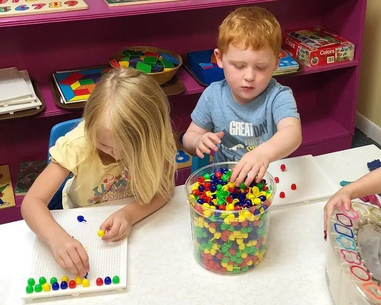 Two children sit at a table placing colorful pegs on white pegboards while a third child's hand reaches toward a container of pegs.