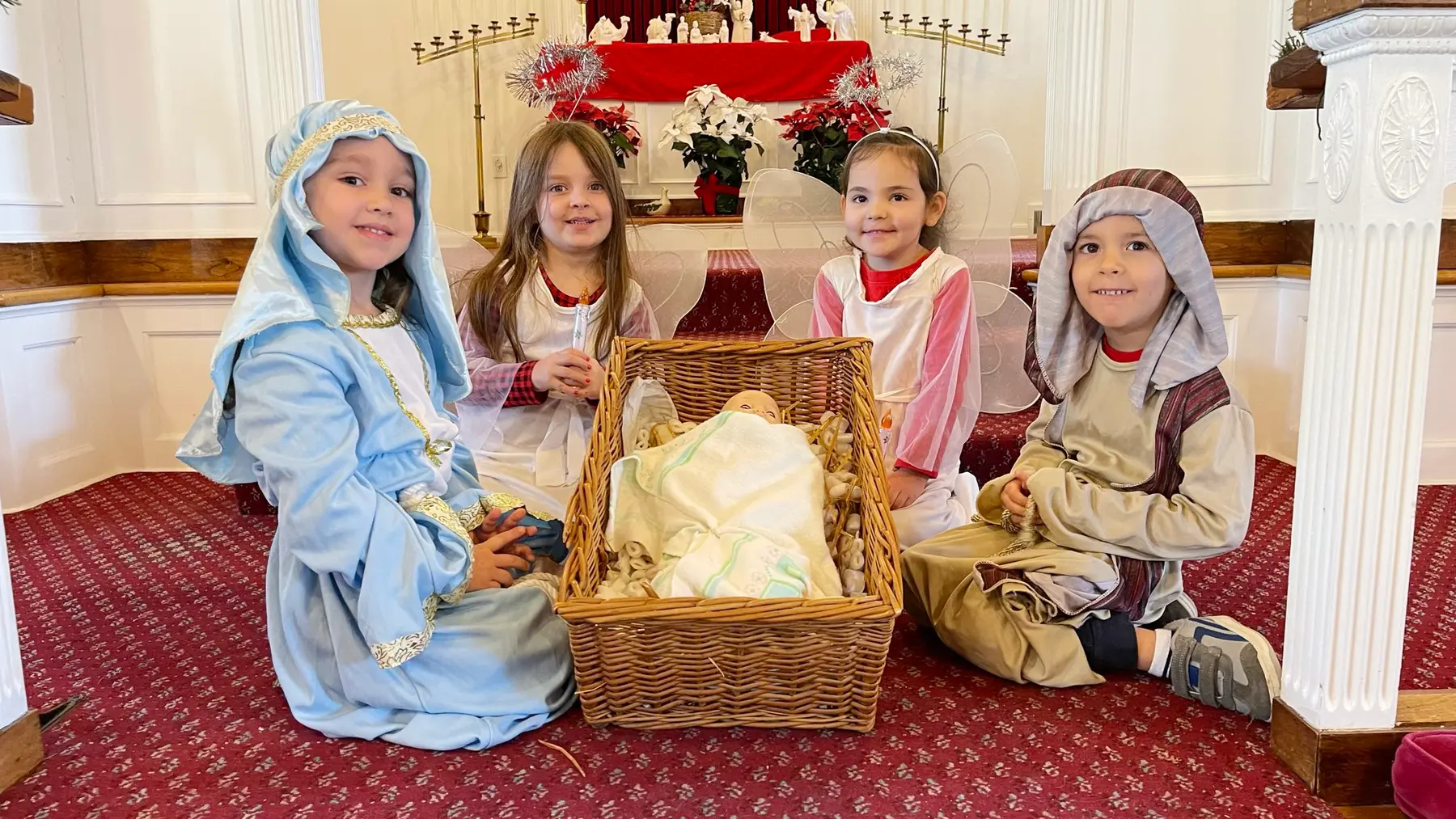 Four children wearing nativity and angel costumes sit on a red patterned carpet around a woven basket holding a doll, with a decorated altar behind them.