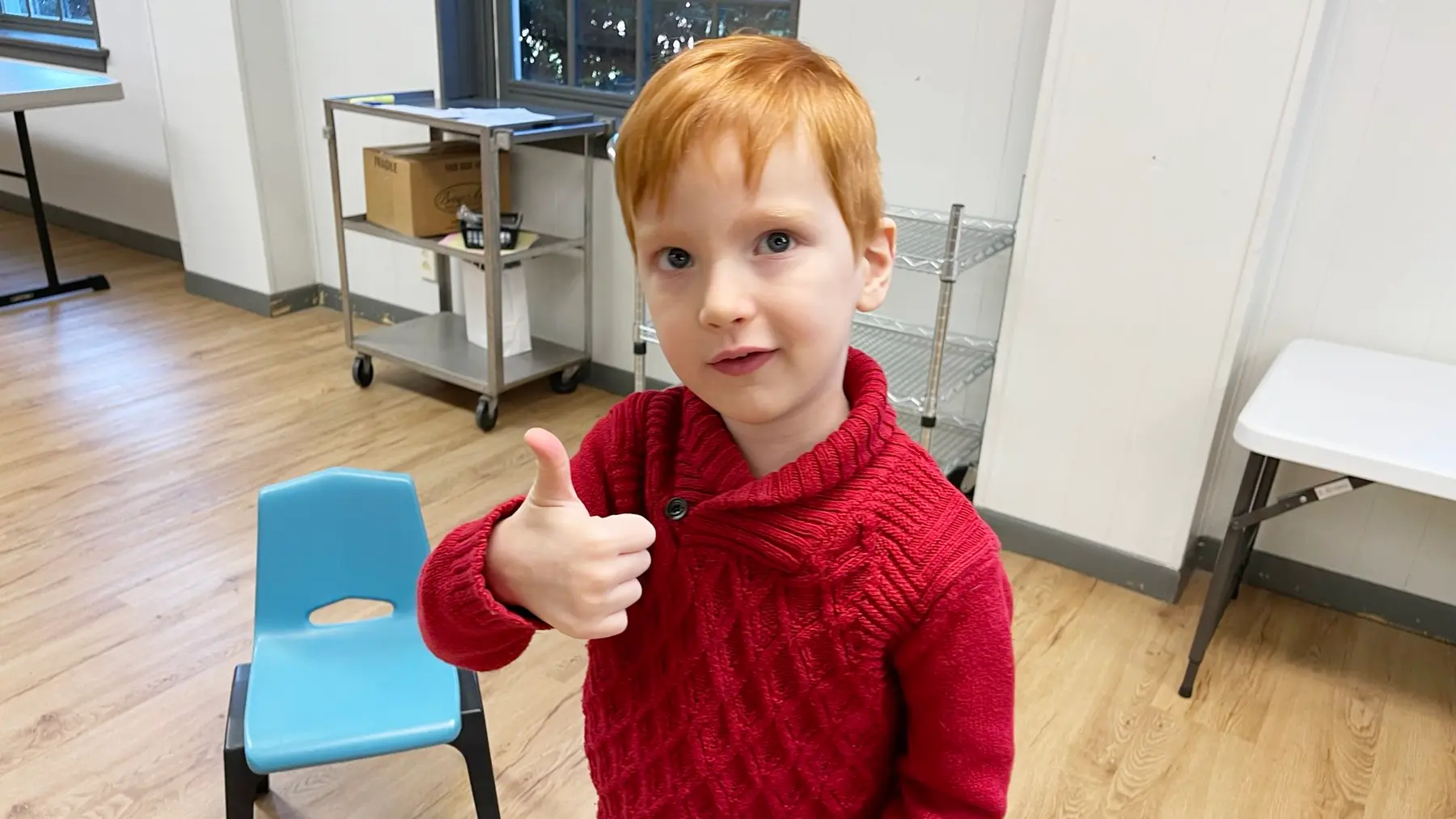 A young boy with red hair wearing a red knit sweater gives a thumbs up while standing in a room with a blue chair, a metal cart, and a white table.