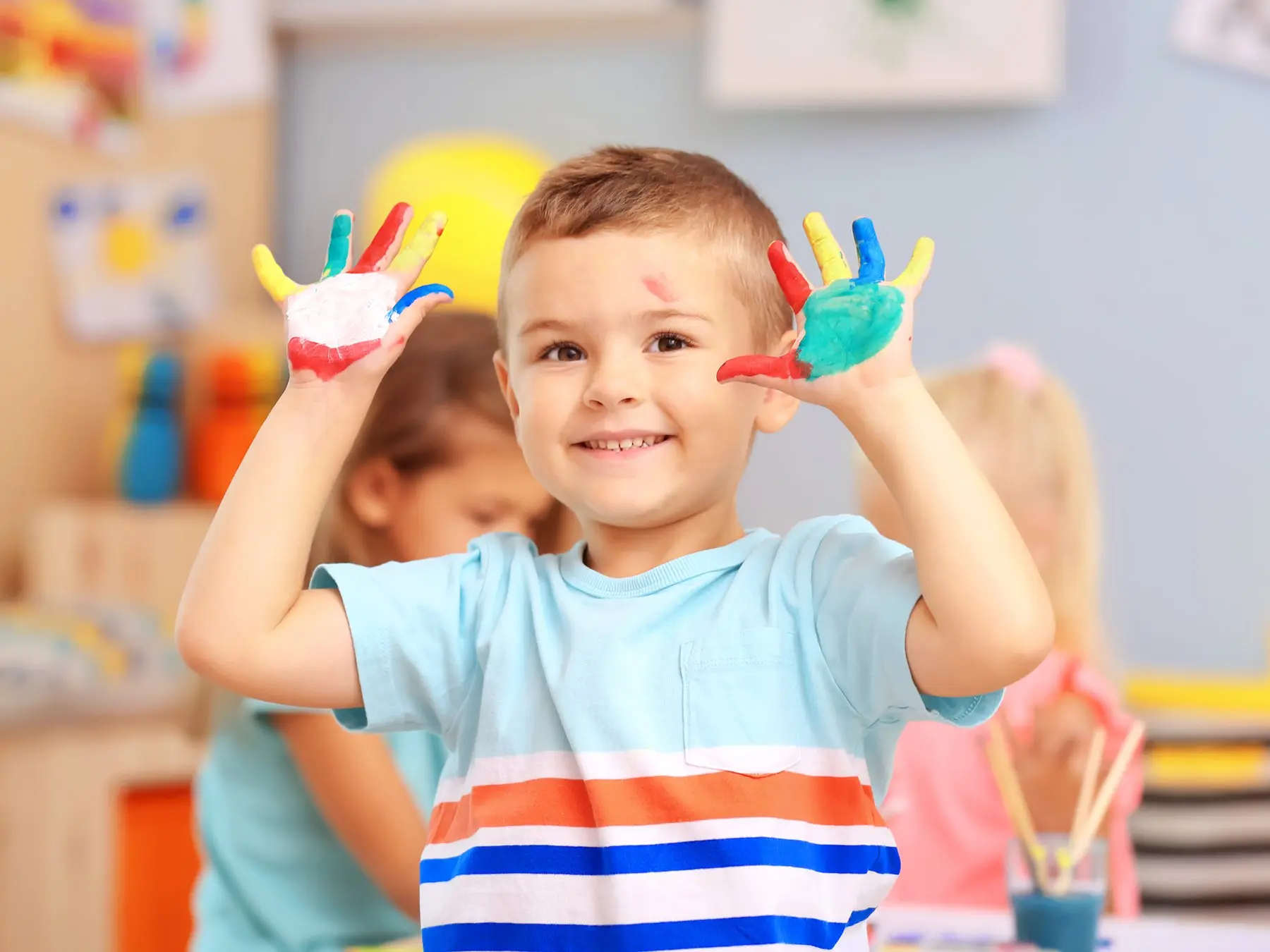 A smiling boy in a striped shirt holds up paint-covered hands in a classroom while two other children sit at a table behind him.