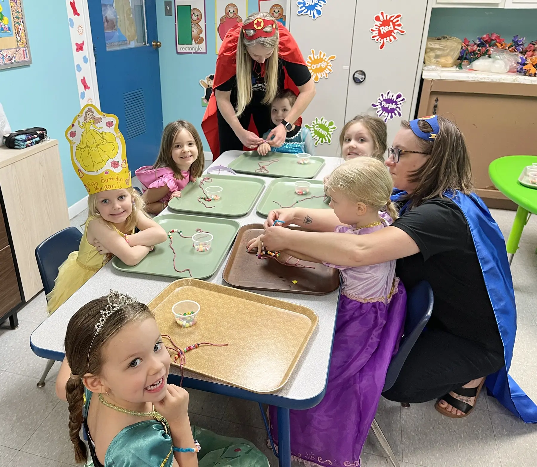 Children in princess costumes and adults in superhero capes sit at tables threading beads onto string on trays in a classroom.