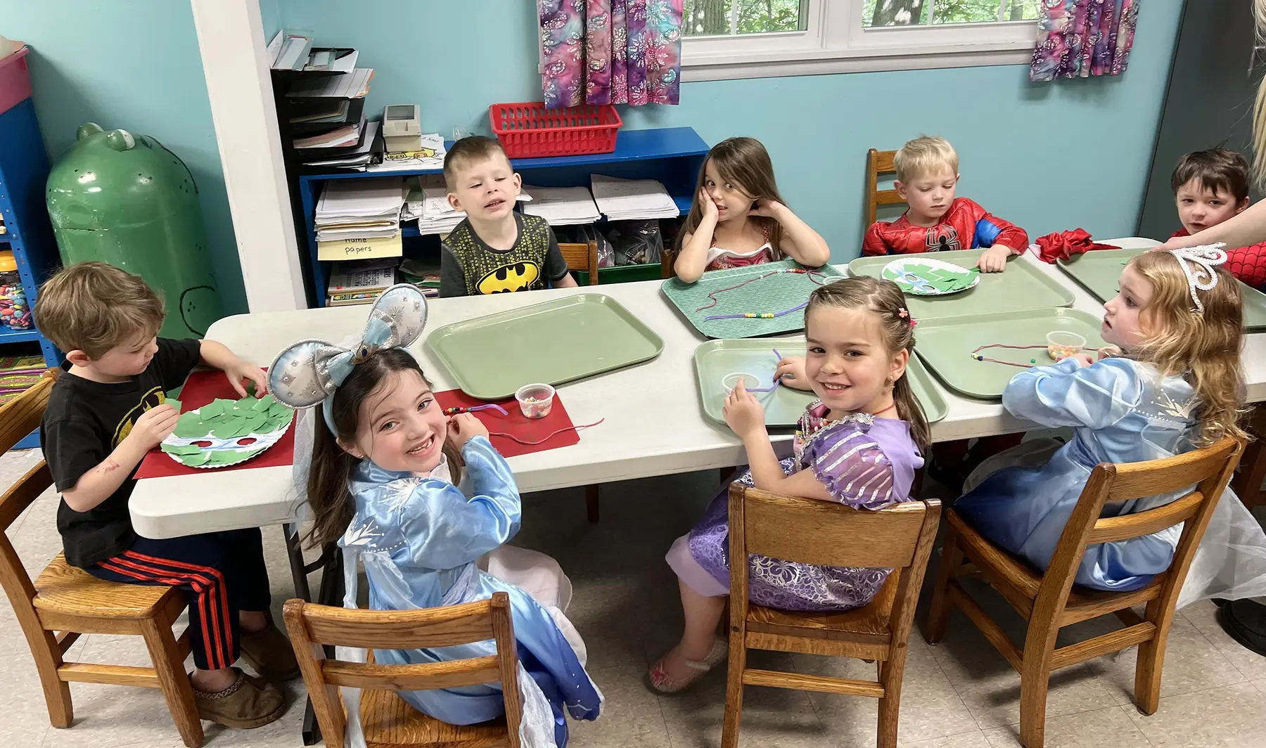 Eight children sit around a white table working on crafts with green trays, paper plates, and string while wearing various costumes and everyday clothes.