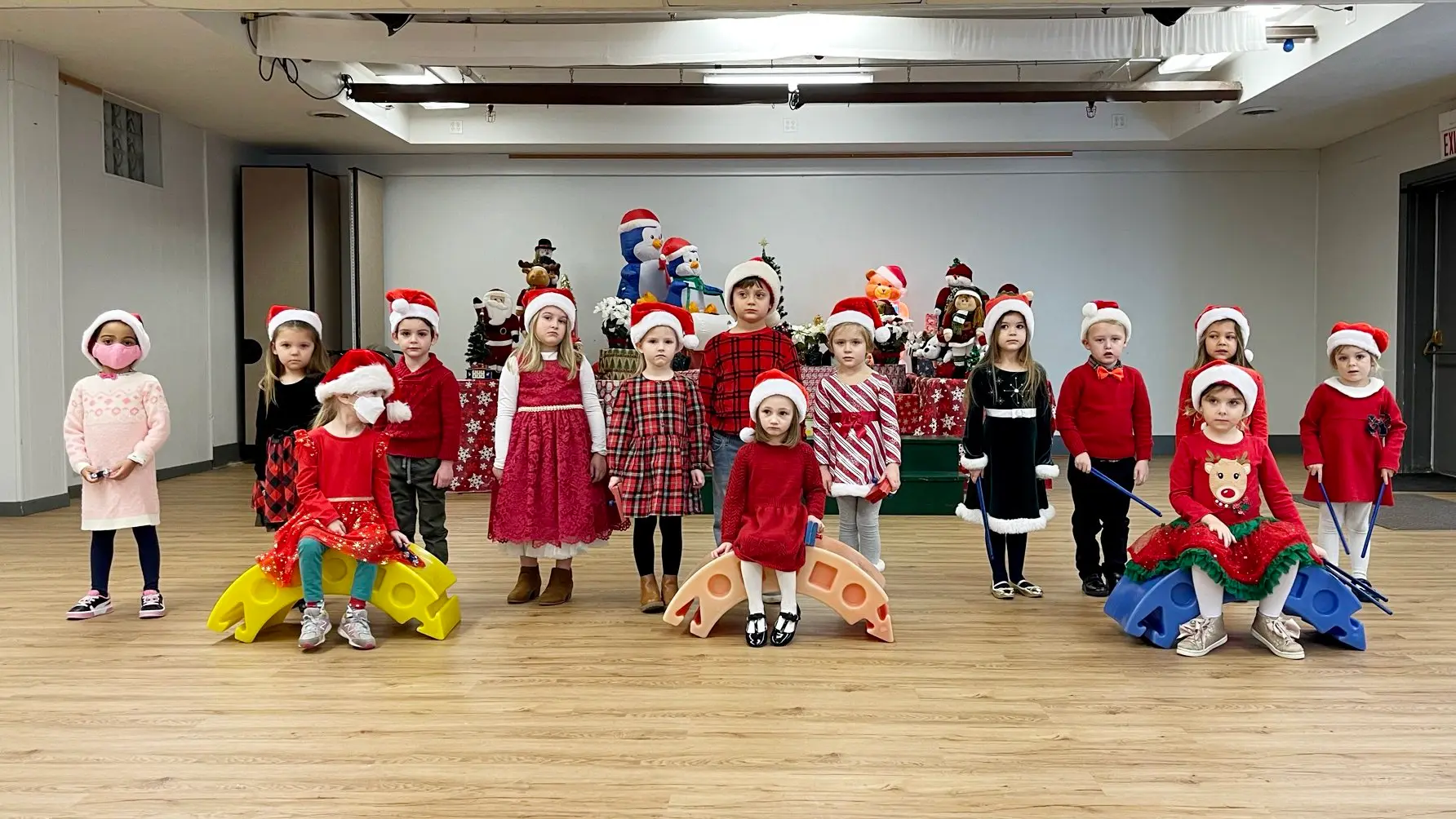 Fourteen children wearing Santa hats and holiday clothing stand and sit on colorful plastic balance boards in a room with a decorated Christmas backdrop.