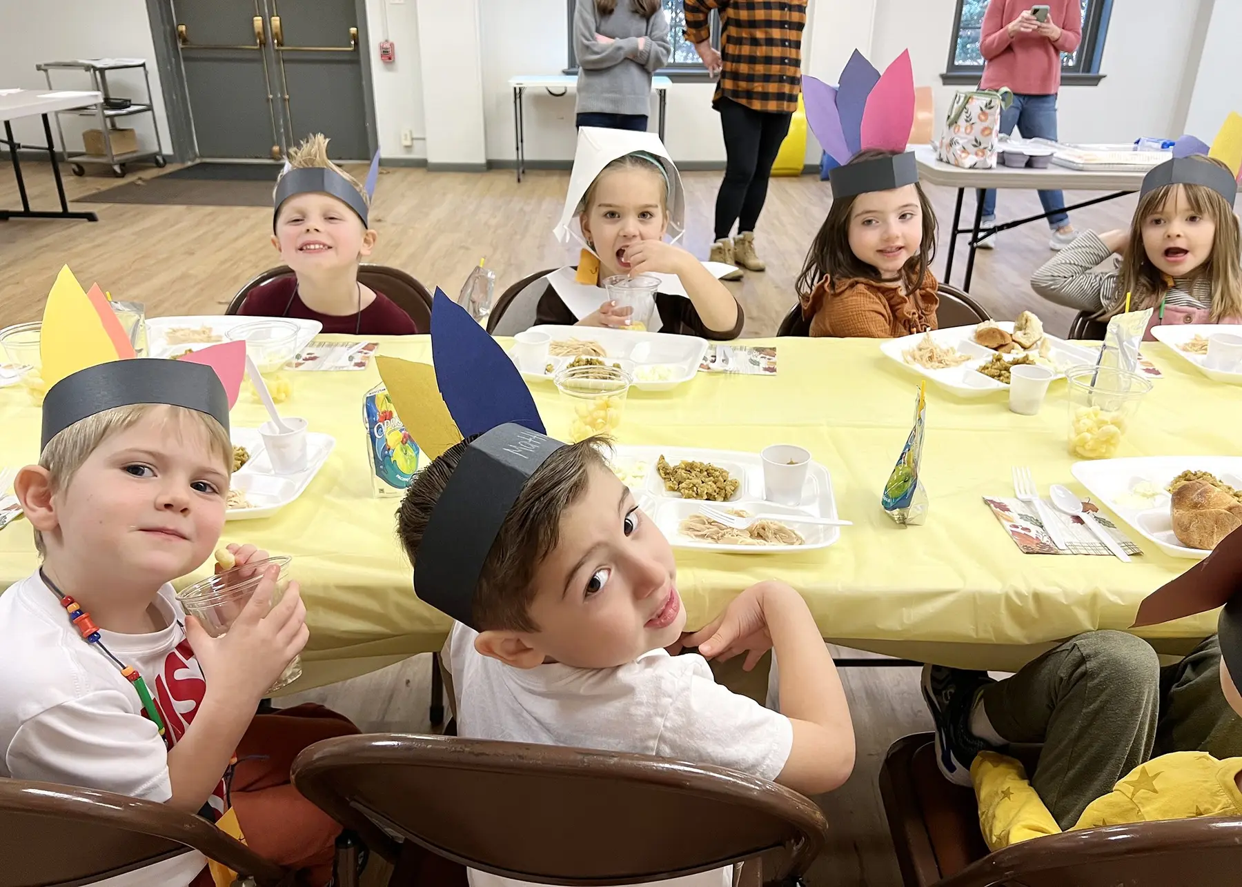 Children wearing paper hats sit at a yellow-covered table with plates of food while adults stand in the background.
