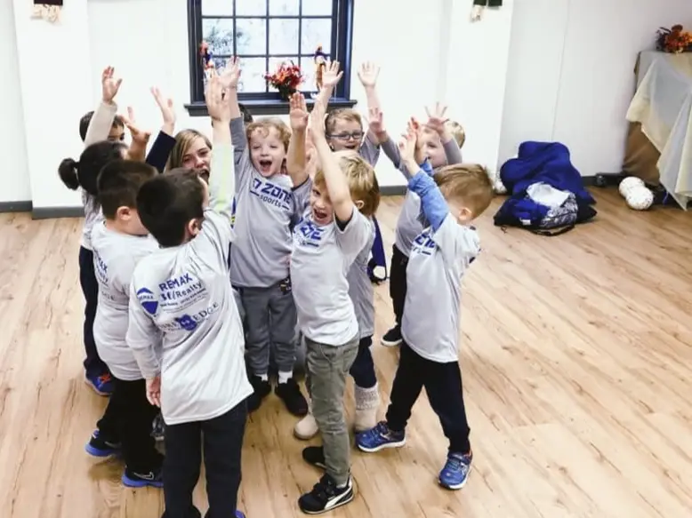 A group of children and one adult stand in a circle with their arms raised in a room with a wooden floor and a window.