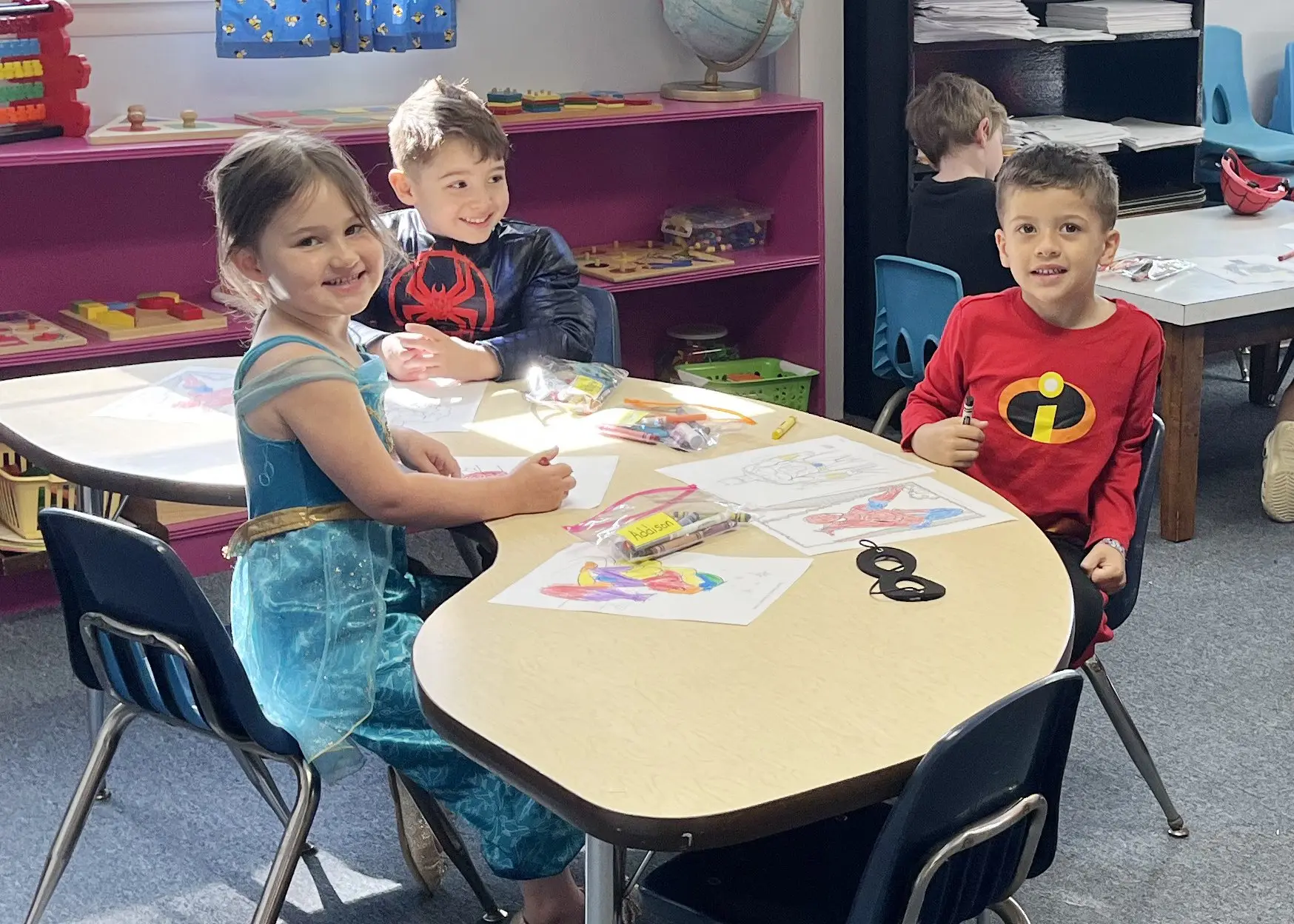 Three children in costumes sit at a table coloring pictures while another child works at a table behind them in a classroom.