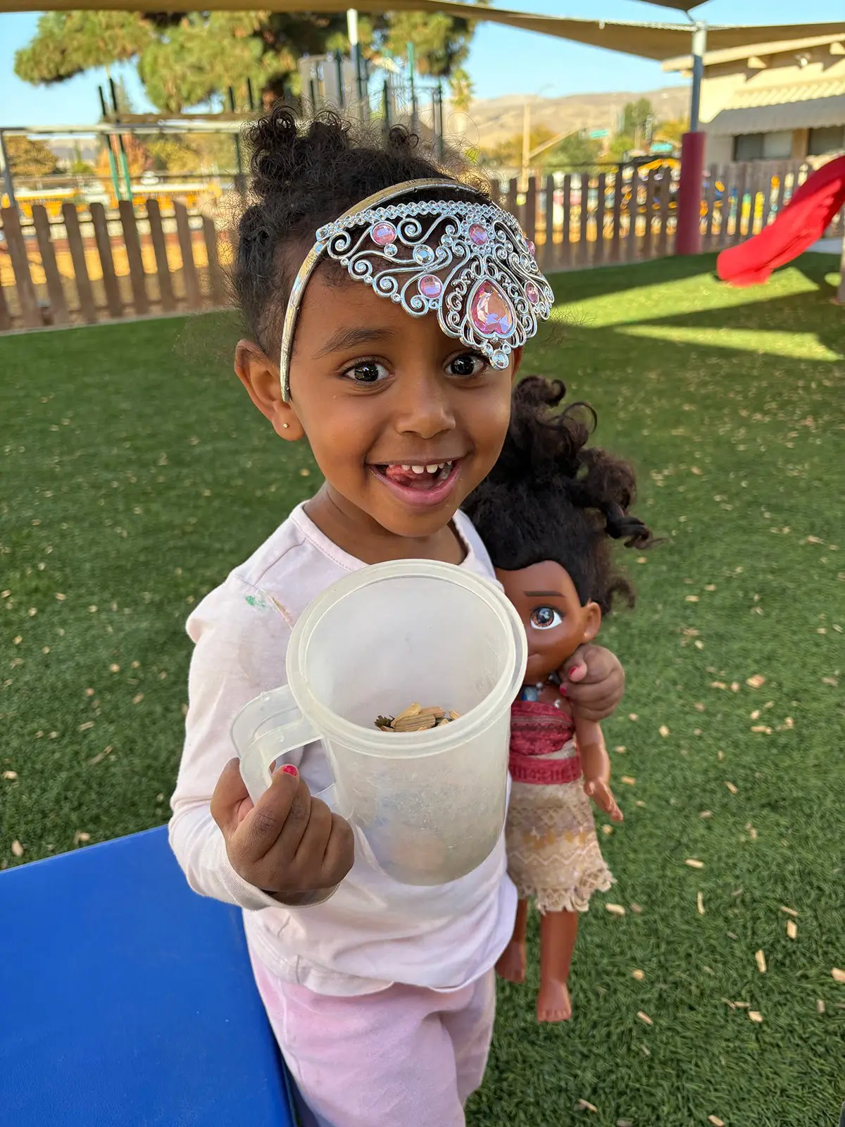 A smiling girl wearing a silver tiara holds a clear plastic pitcher containing small items while carrying a doll on her back.