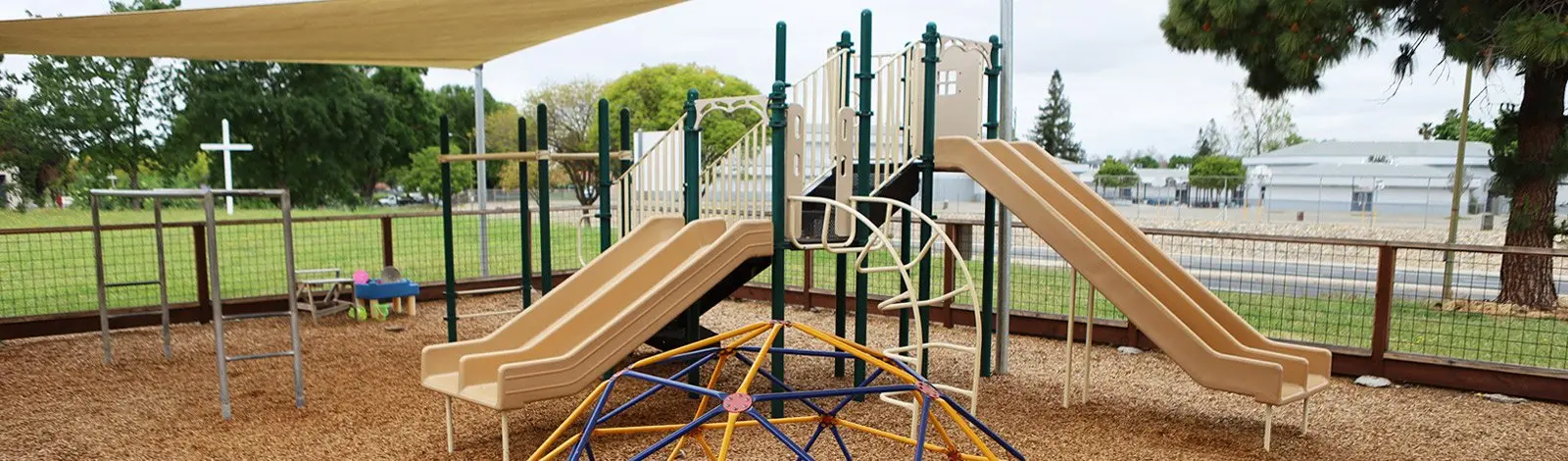 A fenced outdoor playground with a tan canopy, a green and beige play structure with two slides, a climbing dome, swings, and wood-chip ground cover, with no people visible.