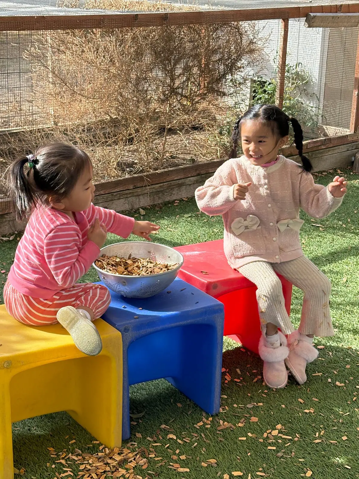 Two girls sit on colorful plastic stools outdoors, one in pink stripes reaching toward a metal bowl of wood chips while the other in a pink cardigan points and smiles.