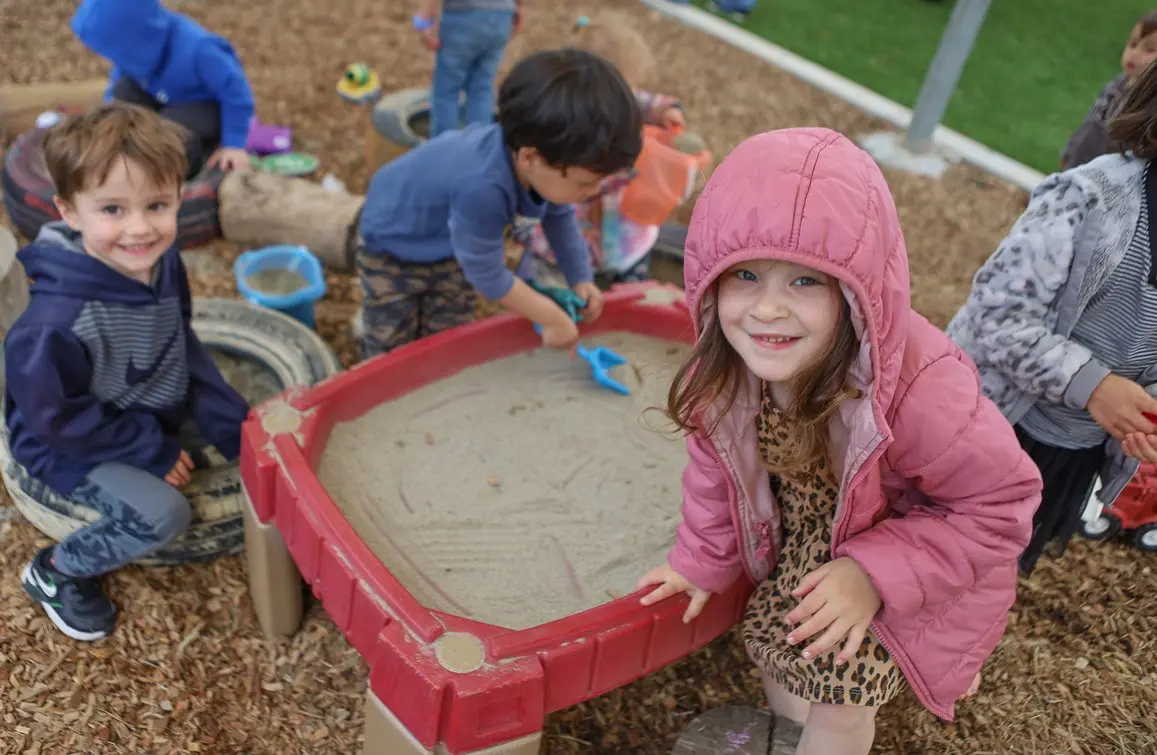Children play with blue shovels in a red sandbox and sit on tires in an outdoor play area covered with wood chips.