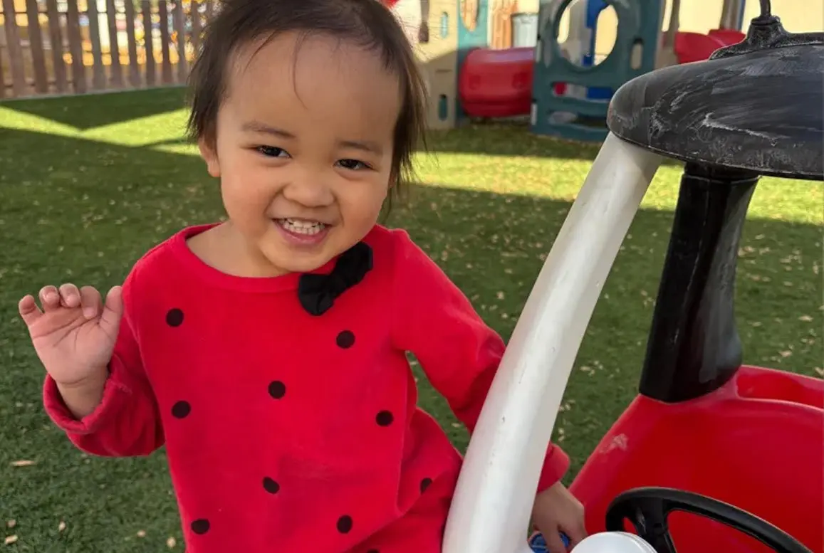 A smiling young child in a red polka-dot dress sits in a red and black toy car on green artificial turf, with playground equipment in the background.