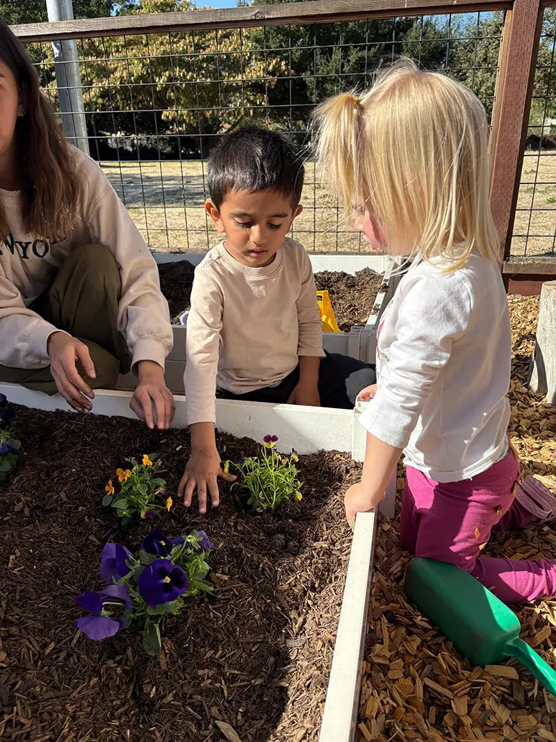 Two young children and an adult kneel beside a raised planter filled with dark mulch and small purple and yellow flowers, with a green watering can resting on the wood-chip ground.