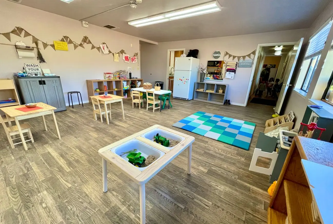 A childcare classroom with small wooden tables and chairs, a sand and toy sensory table, a blue and green checkered rug, storage shelves, and a white refrigerator.